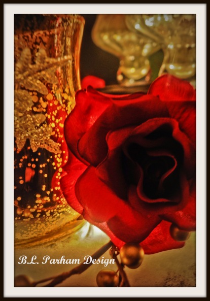 A closeup photograph of a red rose illuminated in the foreground with golden glass candle votives in the background 